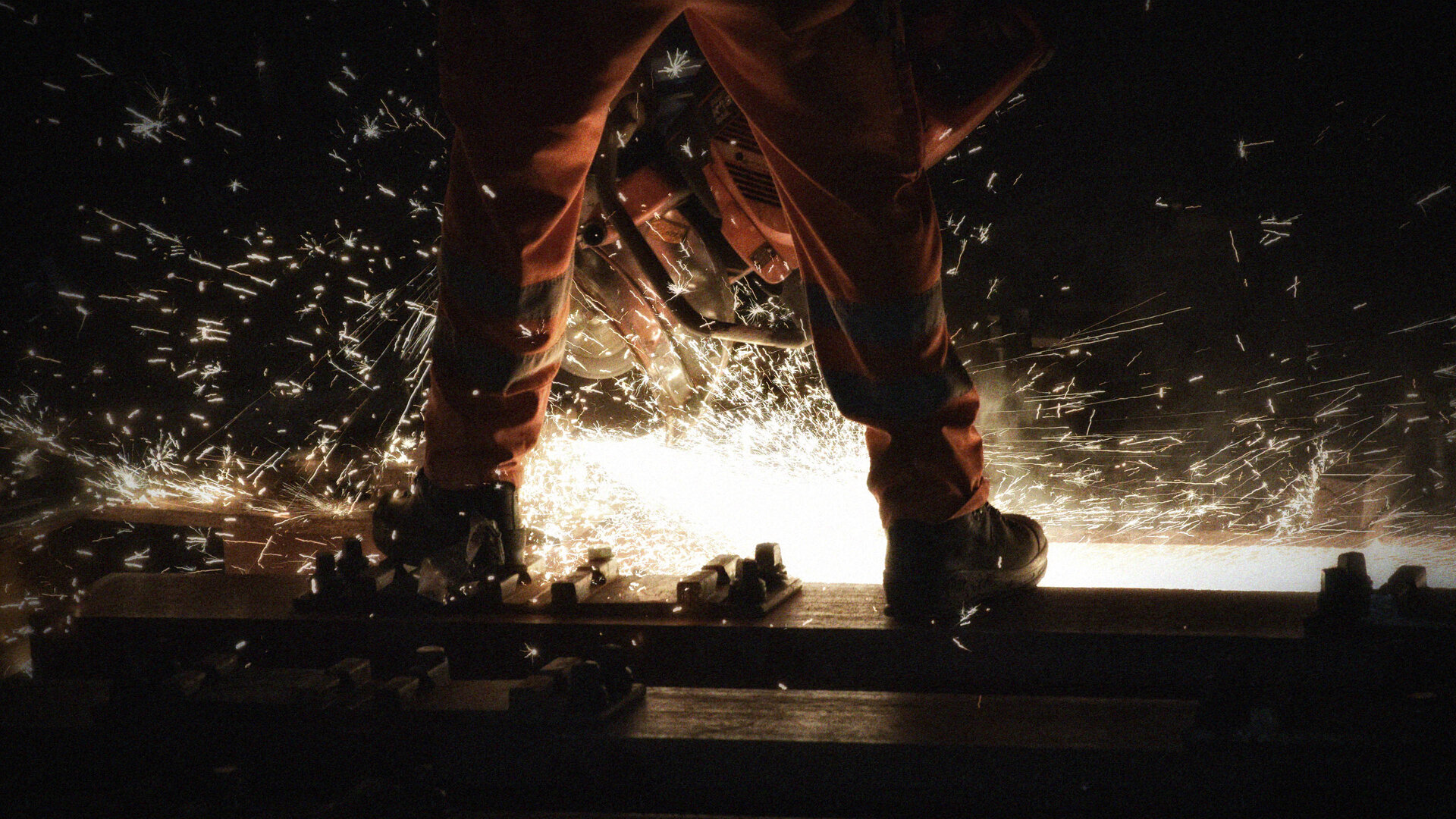 Glühende Schweissfunken fliegen zwischen Beinen einer stehenden Person in Arbeitskleidung.